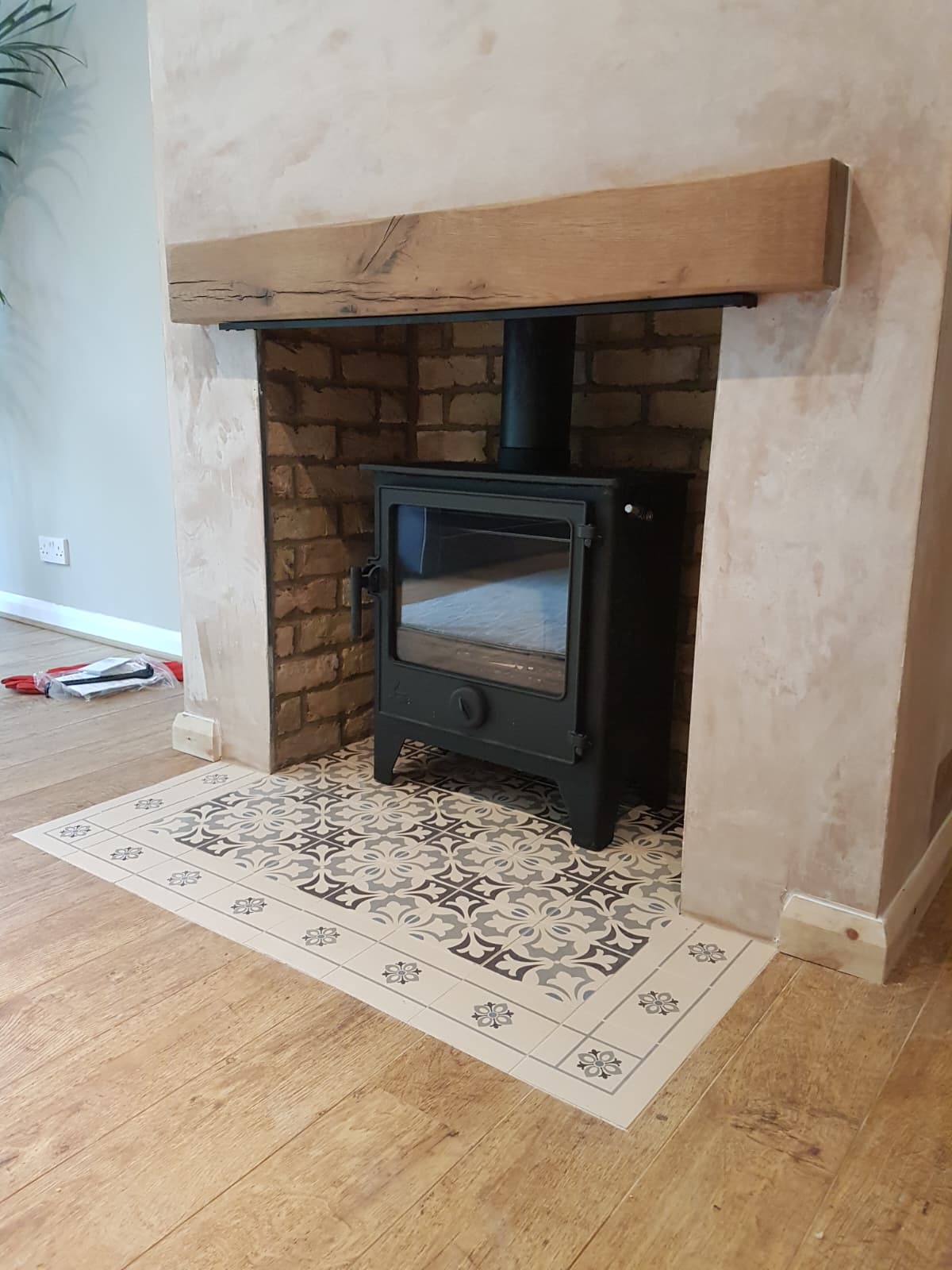 Wood burning stove installed in a rustic brick fireplace with reclaimed oak beam and patterned tile hearth